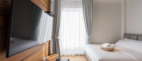 Una habitación de hotel moderna con un televisor de pantalla plana grande montado en la pared, una cama con ropa blanca, una mesita de noche pequeña y una ventana con cortinas de gasa.