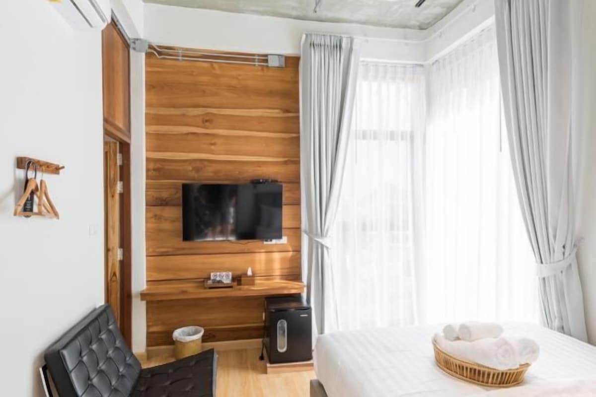 A modern hotel room with a wooden accent wall, a flat-screen TV, a bed with a white comforter, a wicker side table with towels, and a black leather chair.
