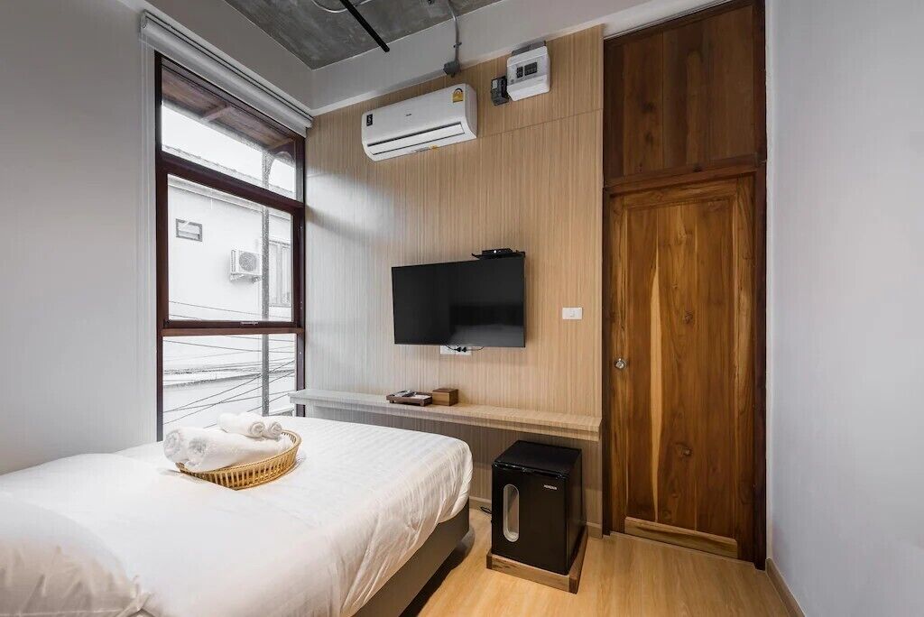 A hotel room with a bed, a TV, an air conditioner, a wooden wardrobe, and a window.
