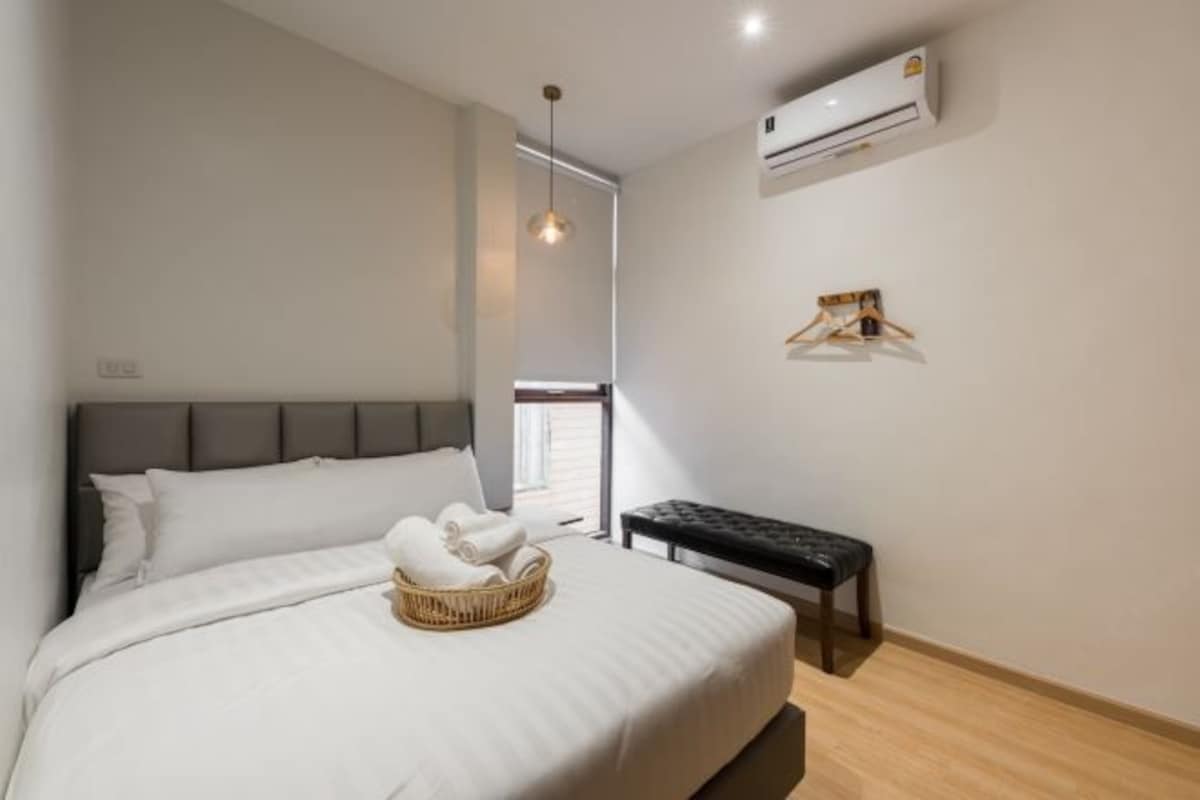 A bedroom with a bed, a bedside table, a hanging light fixture, an air conditioning unit, and a window.