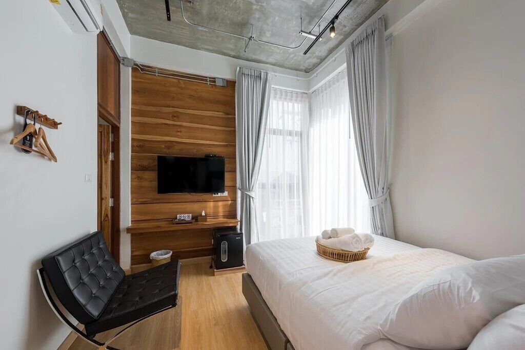 A modern bedroom with a wooden panel wall, a flat-screen TV, a bed with white bedding, a black leather chair, and a small table with a basket.