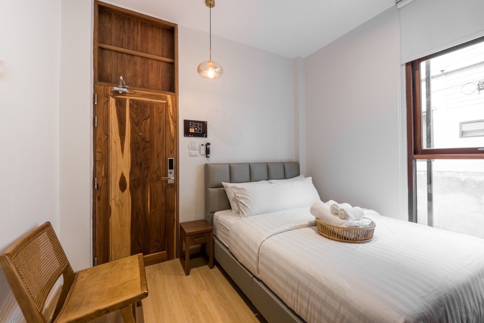 A bedroom with a wooden wardrobe, a bed with white bedding, a small bedside table, a wicker basket, and a hanging light fixture.