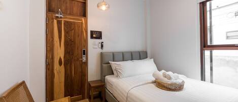 Un dormitorio con un armario de madera, una cama con ropa de cama blanca, una mesita de noche pequeña, una cesta de mimbre y una lámpara colgante.