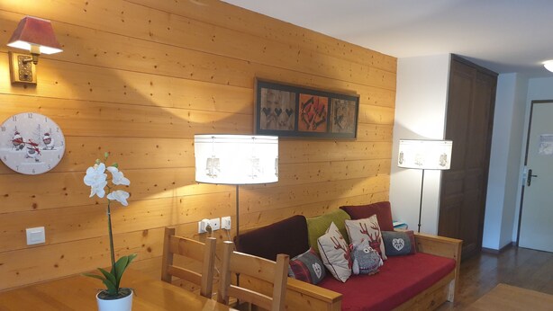 A cozy room with a wooden wall, a wooden table with a potted plant, a sofa with decorative pillows, and a clock. - High quality apartment in the heart of the French Alps - 5 minutes from ski lift (Brides-les-Bains)