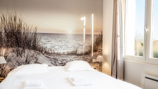 Un dormitorio con una cama grande, ropa de cama blanca, vistas a la playa y una lámpara junto a la ventana.
