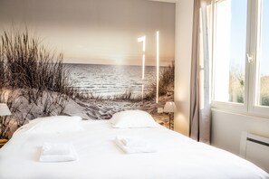 Une chambre avec un grand lit, du linge de lit blanc, une vue sur la plage et une lampe près de la fenêtre.