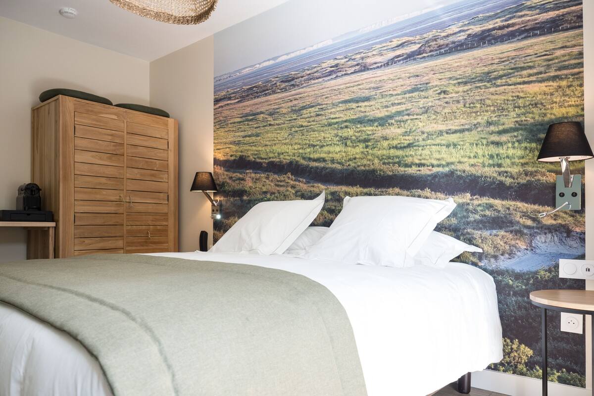 Ein Schlafzimmer mit einem großen Bett, einem Holzschrank, einem Nachttisch, einer Lampe und einer Wandmalerei mit einer Landschaft.