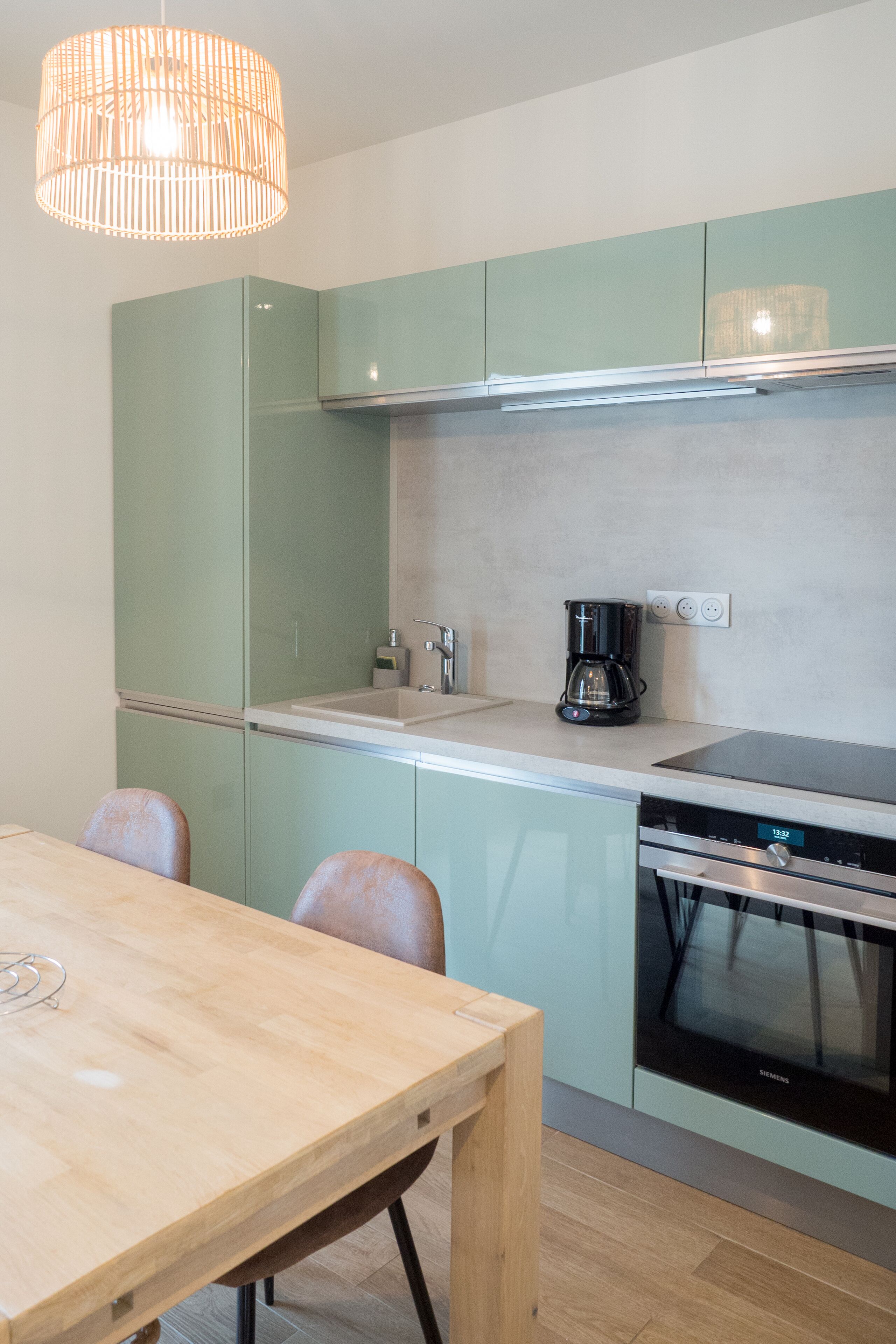 A modern kitchen with light green cabinets, a wooden dining table, and a built-in oven.