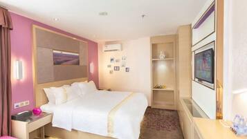 A hotel room with a large bed, a bedside table with a lamp, a small desk, a shelf with decorative items, and a wall-mounted TV.