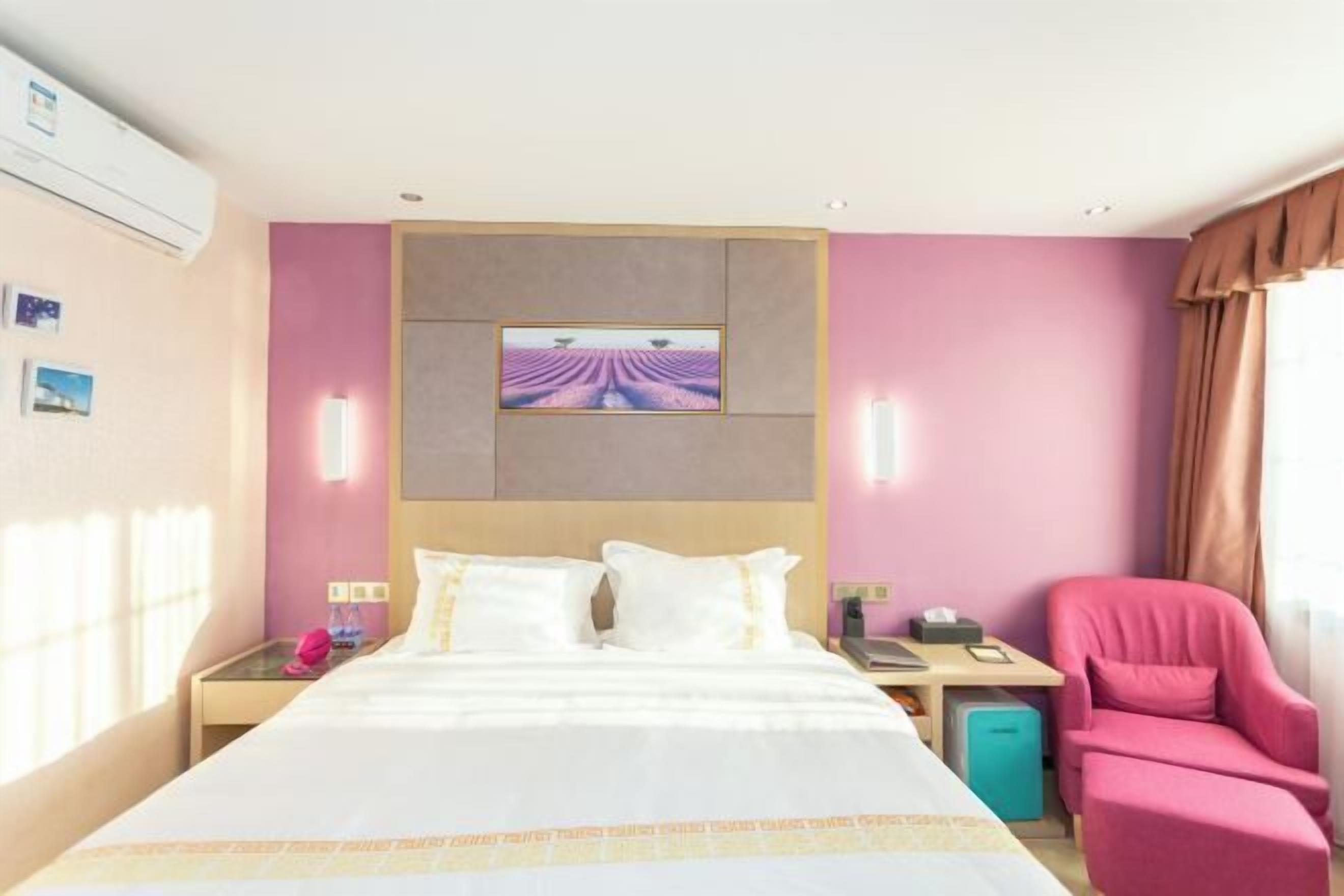 A hotel room with a bed, a pink chair, a desk, and a small table.