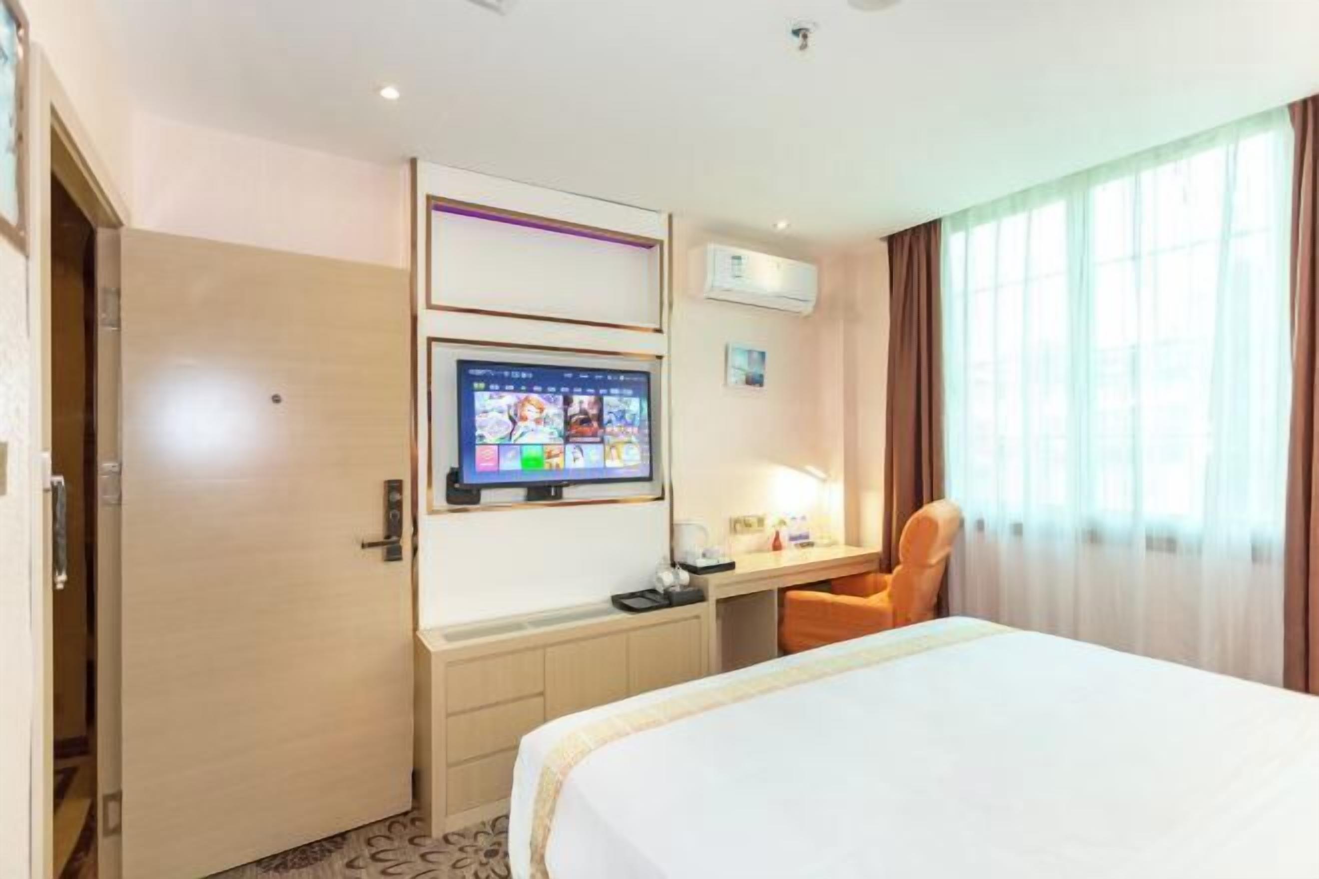 A hotel room with a bed, a desk with a computer monitor, a chair, and a window with curtains.
