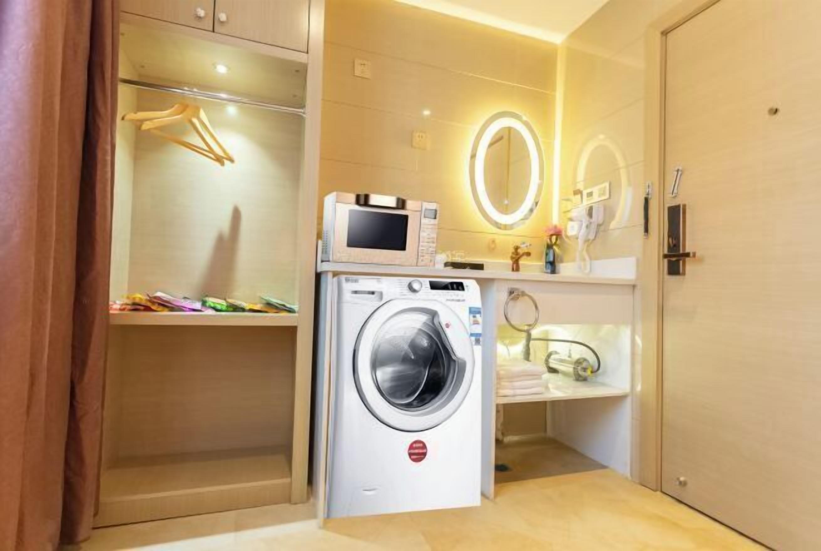 Een compacte hotelkamer met een wasmachine, een magnetron, een make-up tafel met spiegel, en een klein plankje met toiletartikelen.
