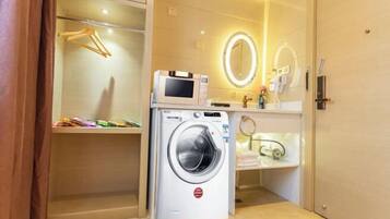 Een compacte hotelkamer met een wasmachine, een magnetron, een make-up tafel met spiegel, en een klein plankje met toiletartikelen.