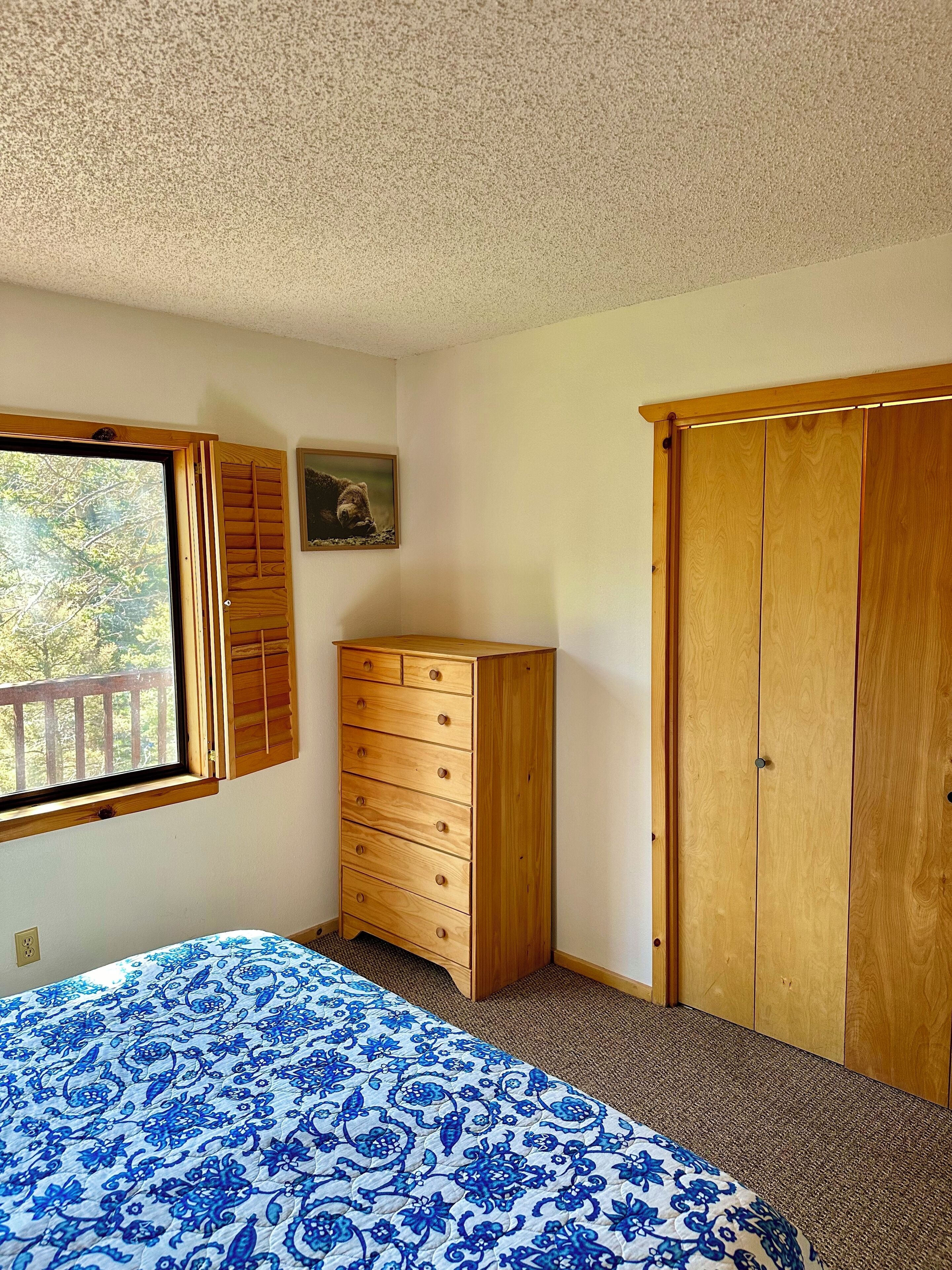 A bedroom with a bed, wooden dresser, wardrobe, window, and a picture on the wall.