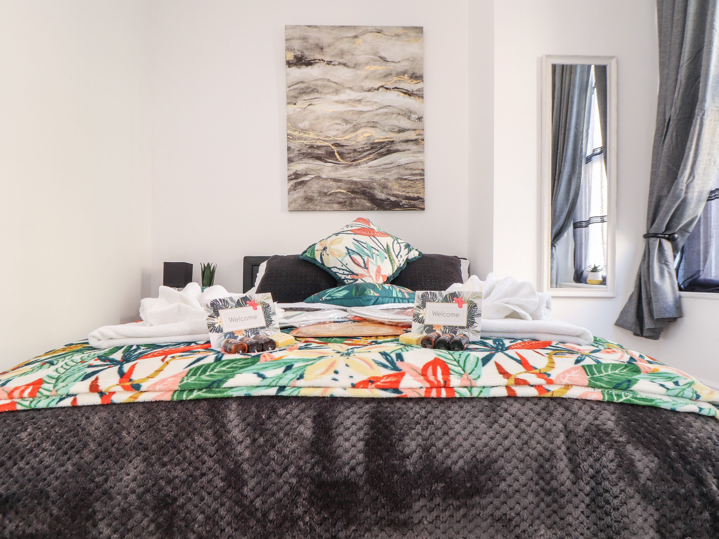 Una cama bien hecha con un edredón floral, una tarjeta de bienvenida y una pequeña planta en la mesita de noche.
