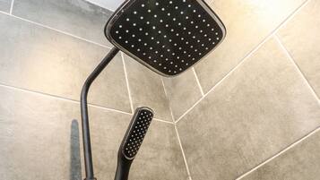 Un cabezal de ducha con diseño cuadrado y múltiples orificios redondos, sujeto a un brazo negro, frente a una pared azulejada.