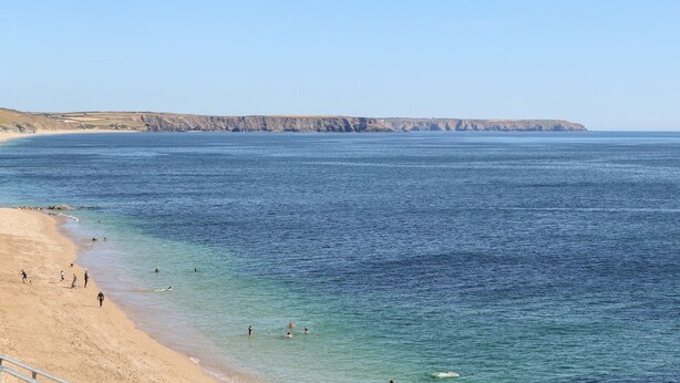 A coastal beach with clear blue water, sandy shore, and rocky cliffs in the background. - The Dynar (Helston)
