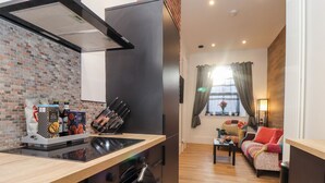 A modern kitchen with a built-in stove, oven, and a brick backsplash. There is a seating area with a sofa and a small table, and a window with curtains. - The Dynar (Helston)