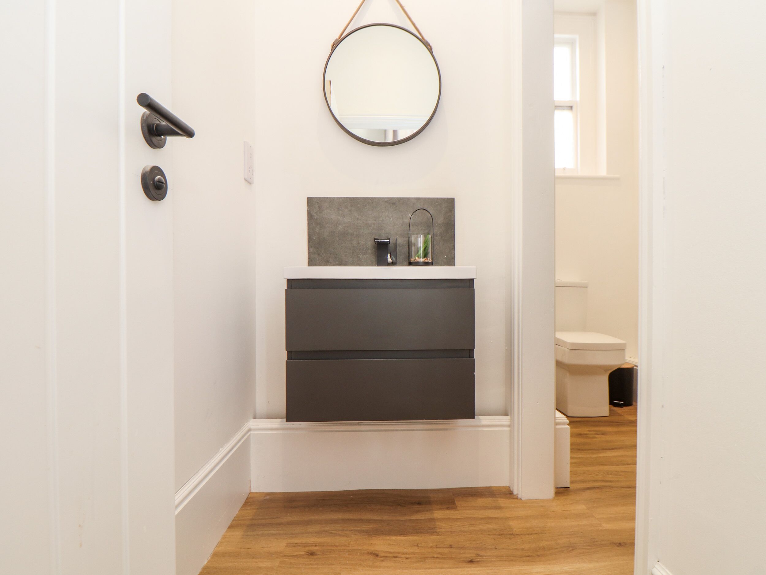 A narrow hallway leading to a bathroom with a white door, a wooden floor, a mirror, and a small sink with a gray countertop.