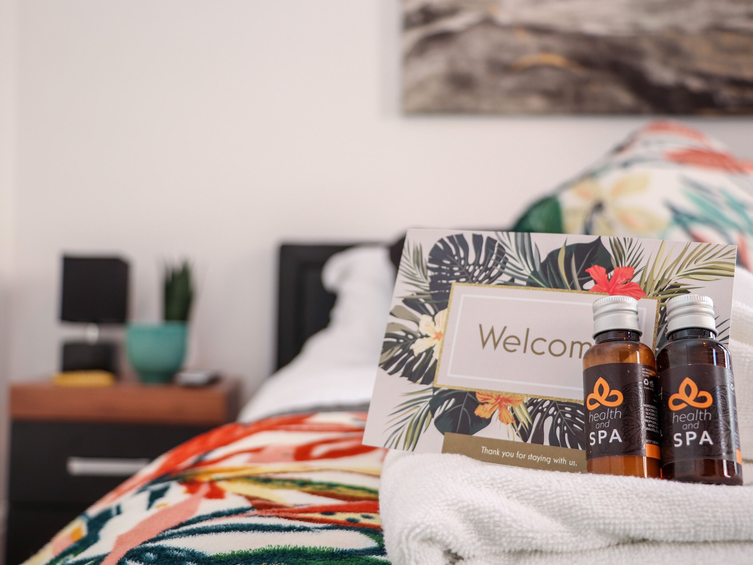 A neatly made bed with a floral-patterned blanket, a welcome card, and two bottles of health and spa products.