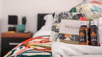 A neatly made bed with a floral-patterned blanket, a welcome card, and two bottles of health and spa products.