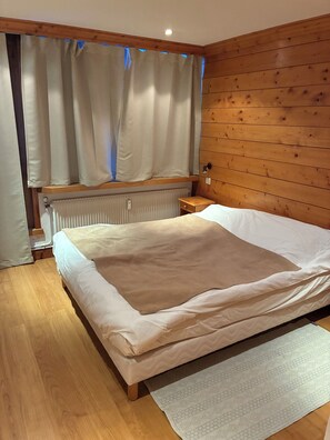 Un dormitorio con cabecera de madera, una cama con manta color beige, una ventana con cortinas, un radiador y una mesita de noche pequeña.