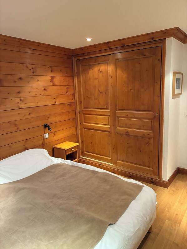 A bedroom with a wooden wardrobe, a bedside table, and a bed with a white and brown blanket.