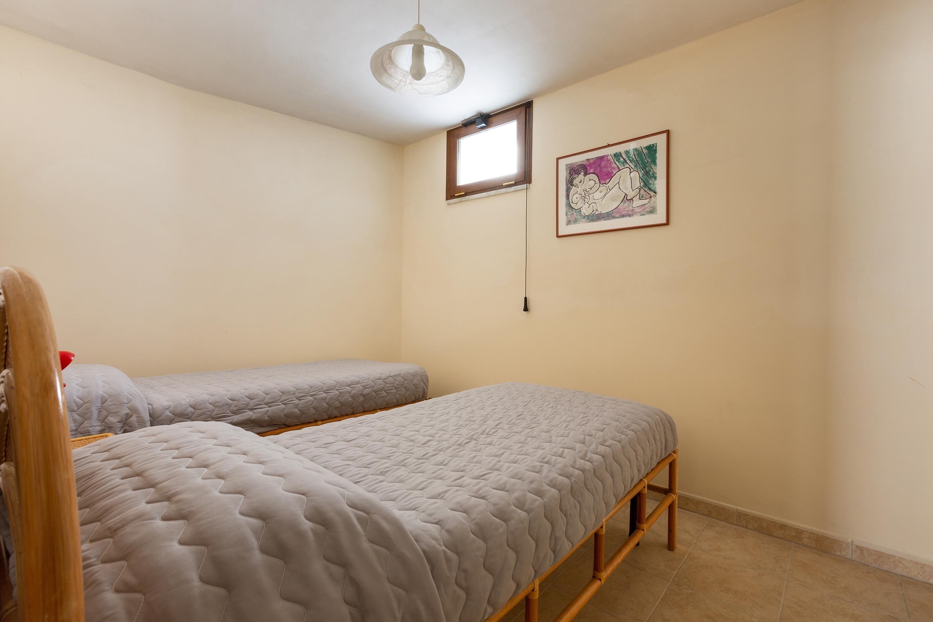 A small, well-lit room with two single beds, a window, and a framed picture on the wall.
