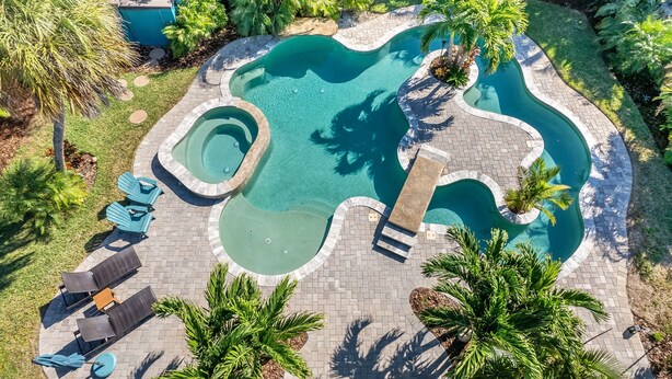 An aerial view of a swimming pool with a wavy design, surrounded by palm trees and a brick patio.