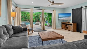A living room with a grey sofa, a wooden coffee table, a TV, and a view of a pool and greenery.