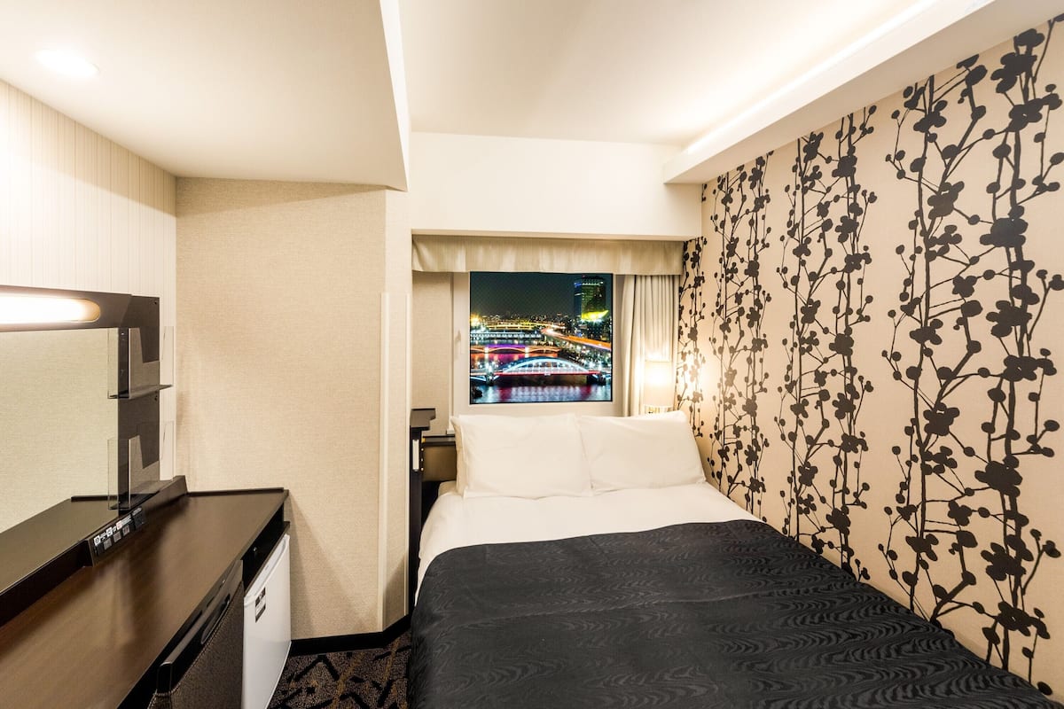 A hotel room with a bed, a desk, a TV displaying a cityscape, and a floral wallpaper.