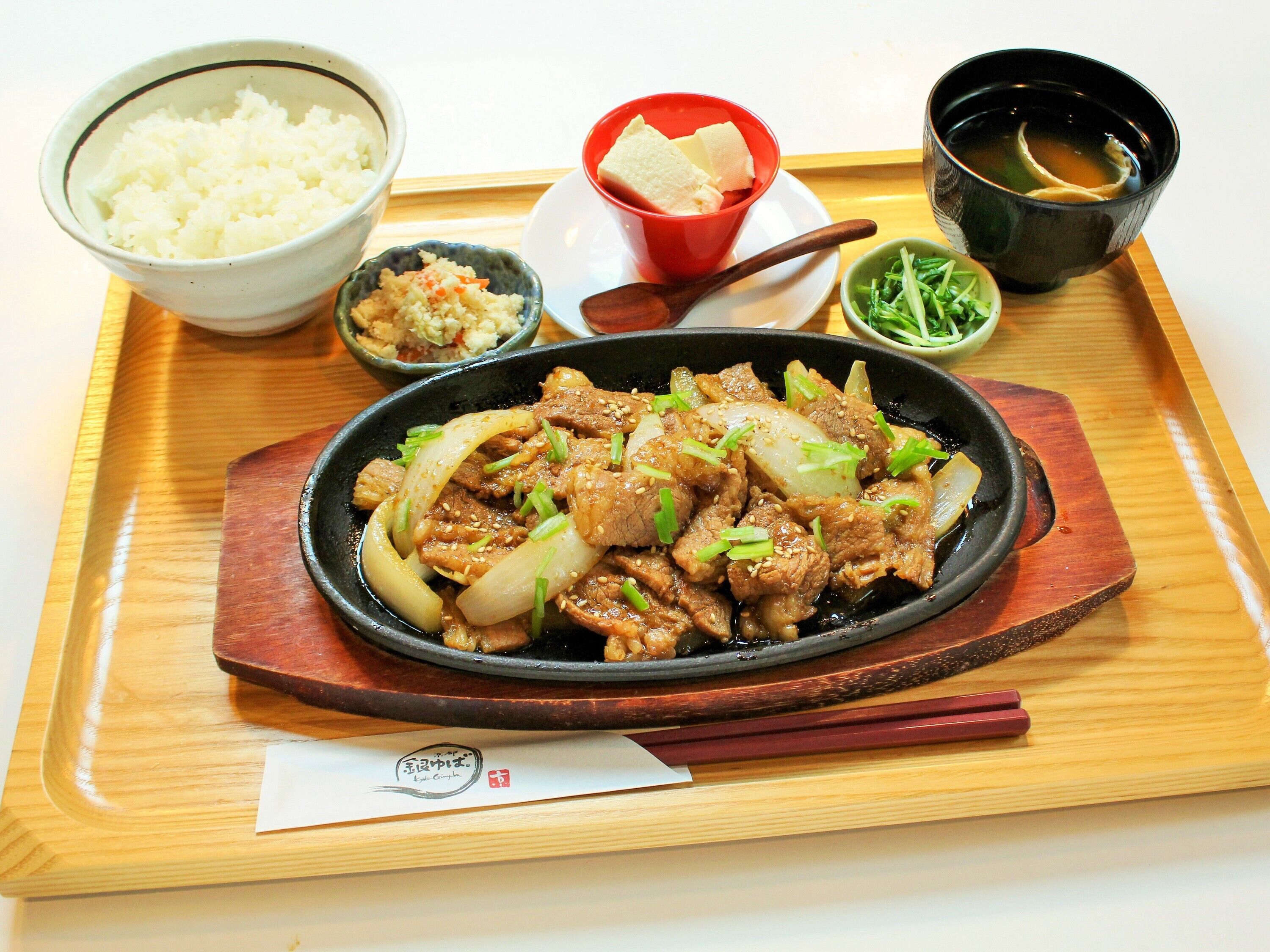 A meal with beef, onions, and green onions, served on a wooden tray with a bowl of rice, a small bowl of sauce, and a cup of tea.
