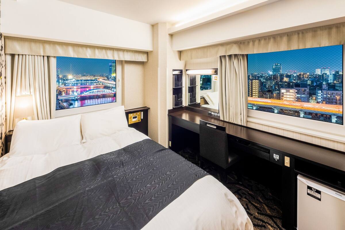 A hotel room with a bed, a desk, a mini-fridge, and a large window with a city view.