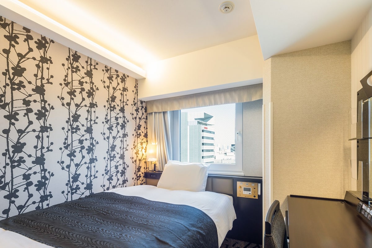 A hotel room with a large bed, a desk, a lamp, and a window with a city view.