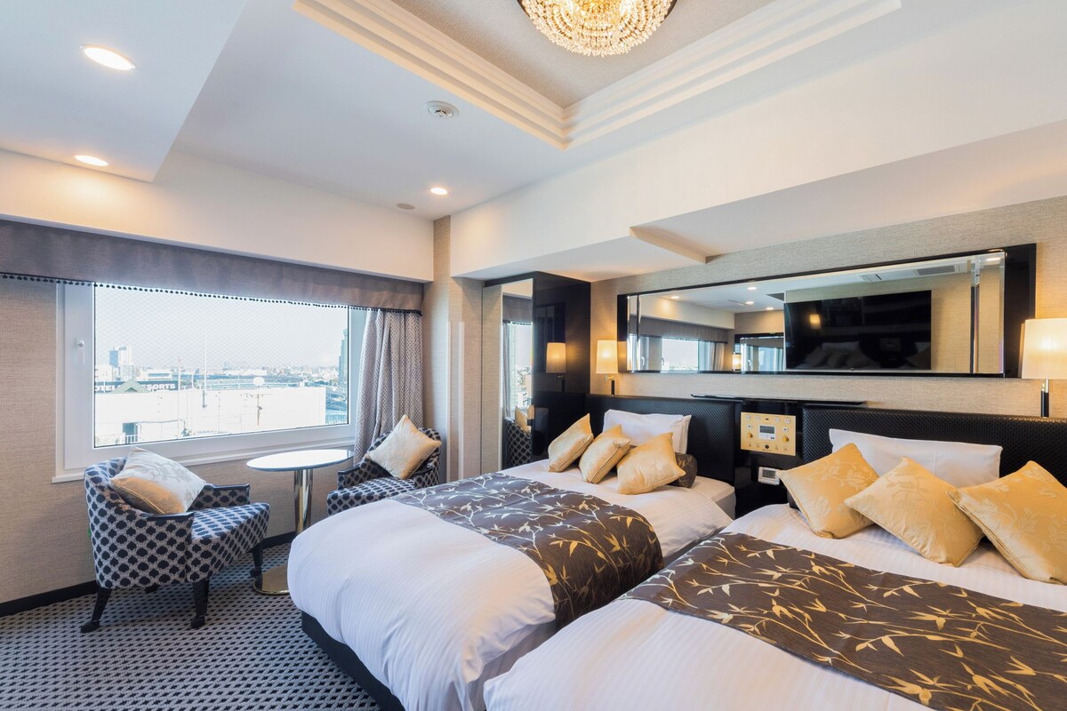 A hotel room with two beds, a seating area with a table, a large mirror, and a view of the city.