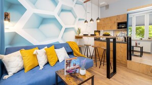 A modern living room with a blue sofa, yellow and white cushions, a wooden coffee table, and a magazine. A kitchen with wooden cabinets and a window is visible in the background. - Comfortable Studio in Cracow | Place to Work | Close to Market Square (Kraków)