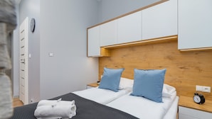 A modern hotel room with a wooden headboard, two beds with white linens and blue pillows, a black textured bedspread, a white towel on the bed, a nightstand with a clock, and a wall-mounted light. - Comfortable Studio in Cracow | Place to Work | Close to Market Square (Kraków)
