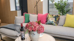 Un salon moderne avec un canapé, des coussins, une table ronde ornée d’un vase de fleurs, des verres à vin et une bouteille.