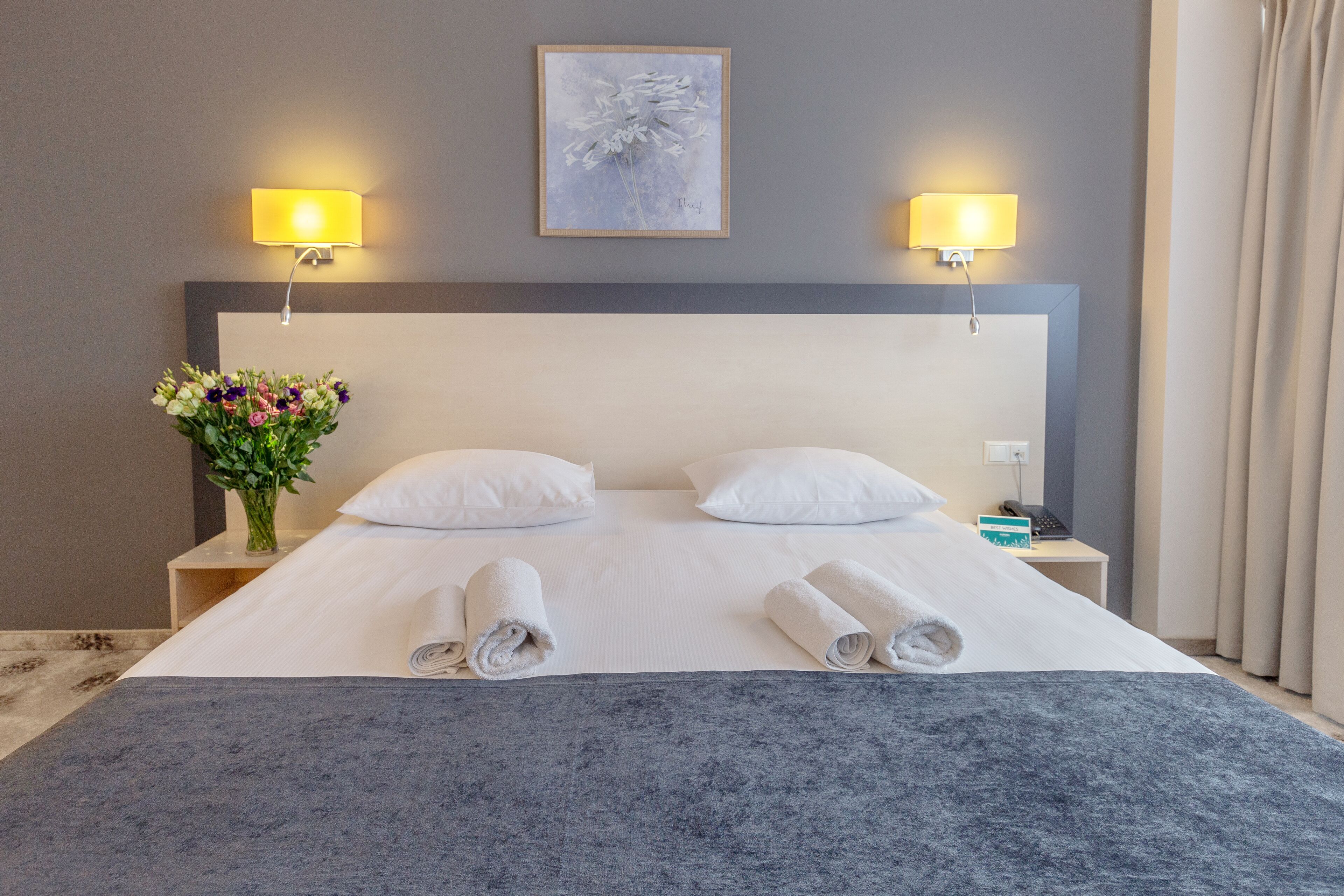 A hotel room with a large bed, two bedside lamps, a vase of flowers, and a painting on the wall.