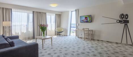 A hotel room with a sofa, a small table with a vase of flowers, a TV on the wall, and a tripod.