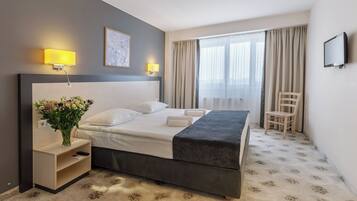 A hotel room with a bed, bedside lamps, a TV, a chair, and a window with curtains.