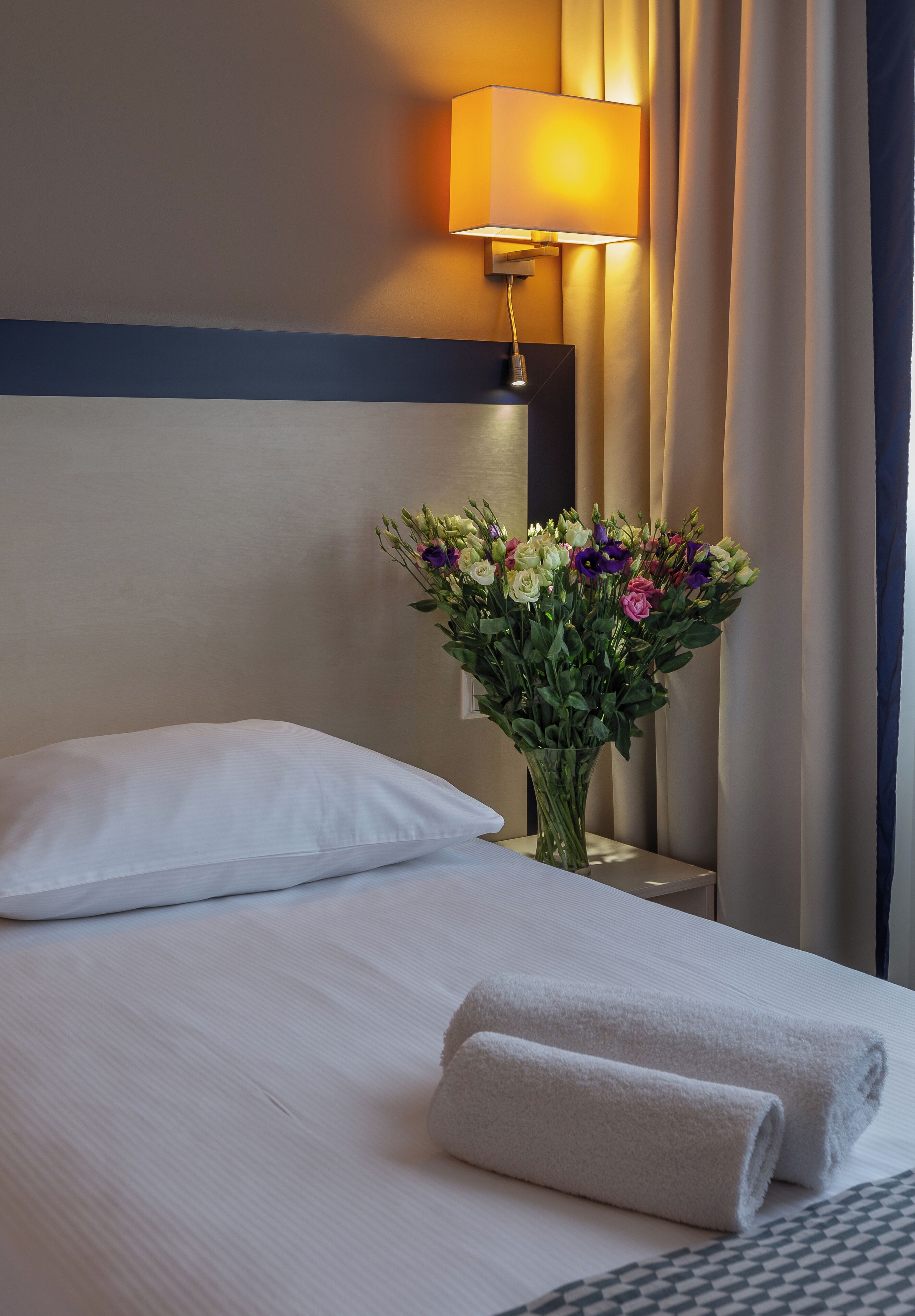 Una cama bien hecha con una almohada blanca, un florero con flores y una toalla enrollada en una habitación de hotel.
