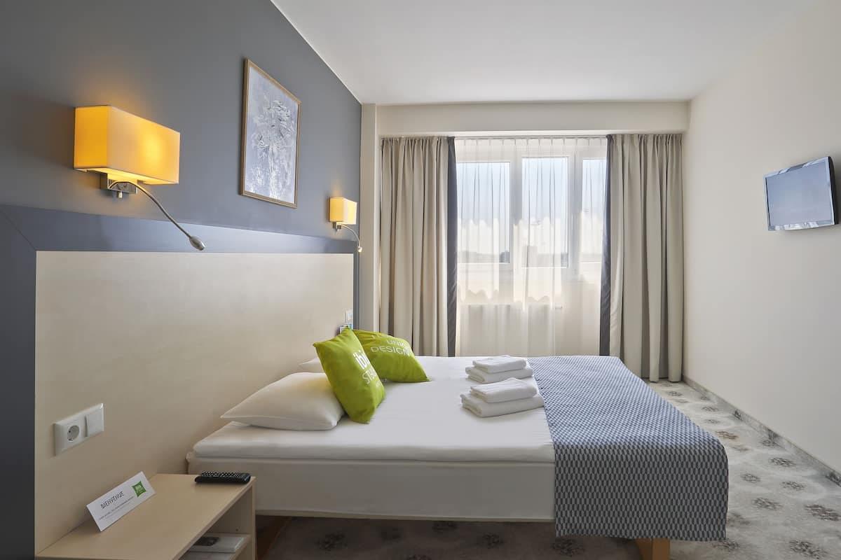 Ein Hotelzimmer mit Bett, Nachttisch, einem an der Wand befestigten Fernseher und einem Fenster mit Vorhängen.