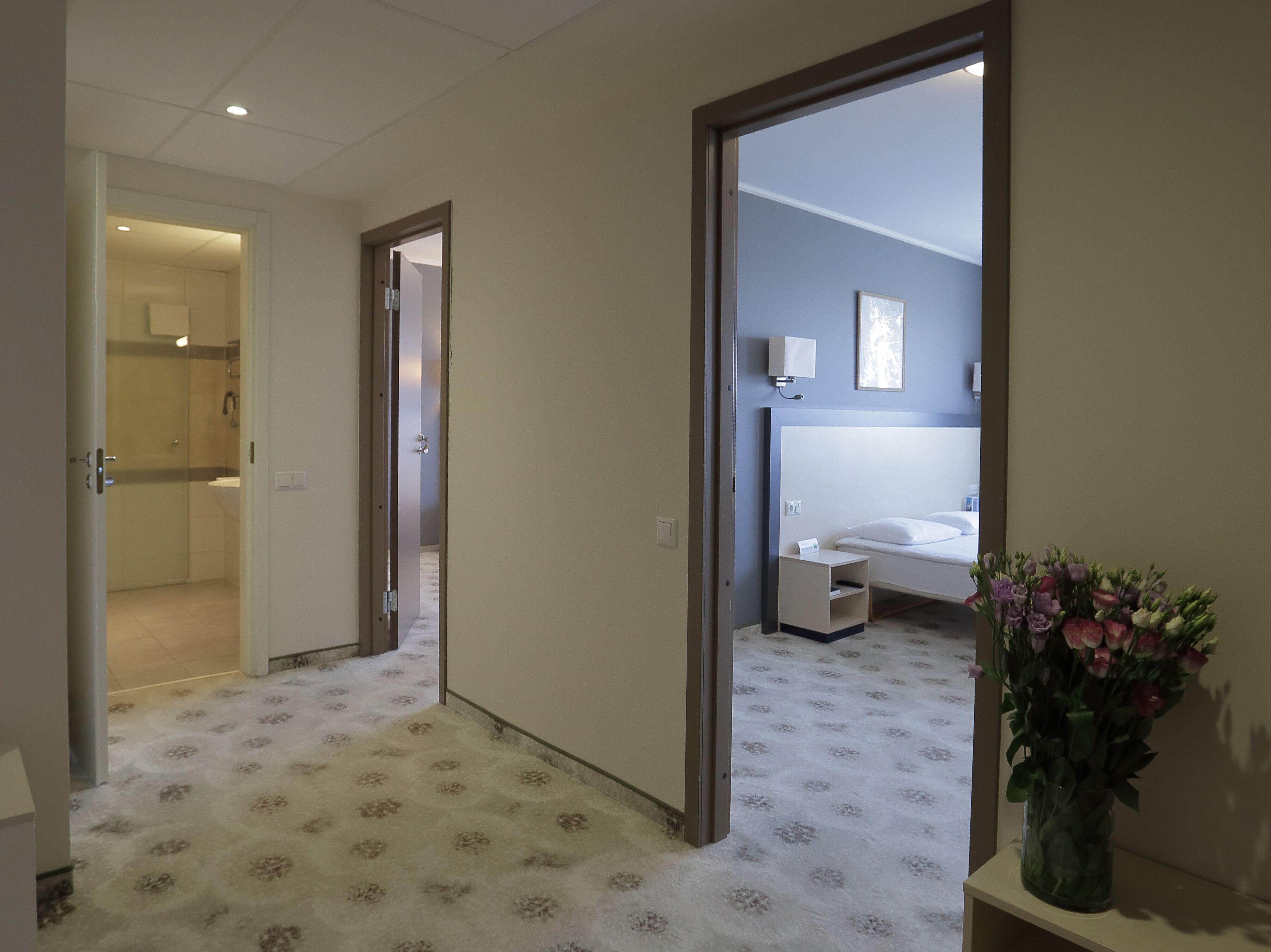 A hotel corridor with a patterned carpet, a bed in the background, and a vase of flowers on a table.