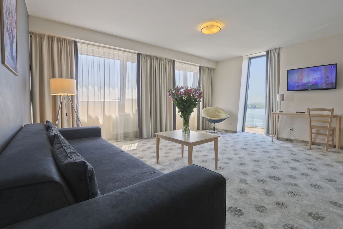 Ein Hotelzimmer mit einer Couch, einem kleinen Tisch mit einer Vase, einem Sessel, einem Schreibtisch mit Fernseher und großen Fenstern mit Vorhängen.