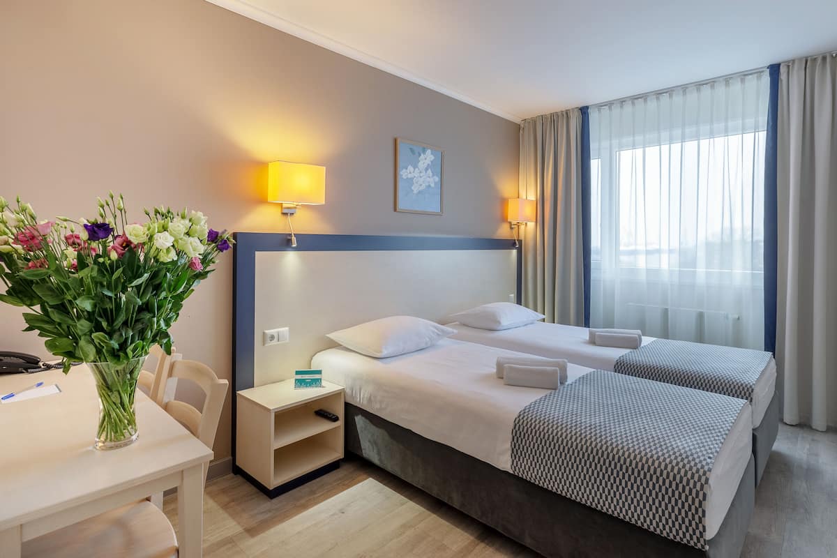 Ein Hotelzimmer mit zwei Betten, einem Schreibtisch, einer Blumenvase und einem Fenster mit Vorhängen.