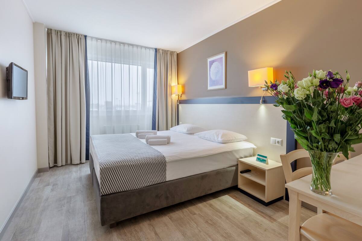 Ein Hotelzimmer mit Bett, Nachttisch, Lampe und Blumen in einer Vase.