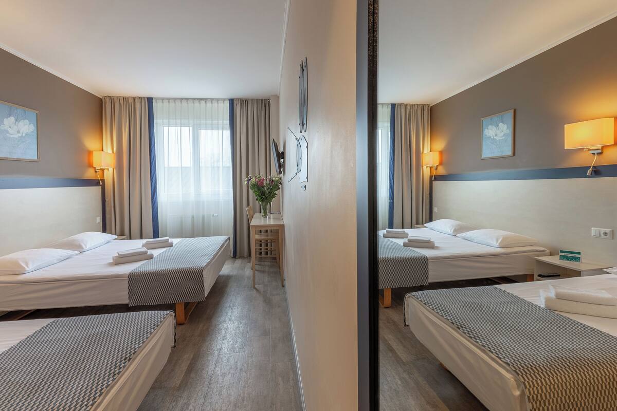 Ein Hotelzimmer mit zwei Einzelbetten, einem kleinen Tisch mit Blumen und einem Spiegel, der den Raum reflektiert.