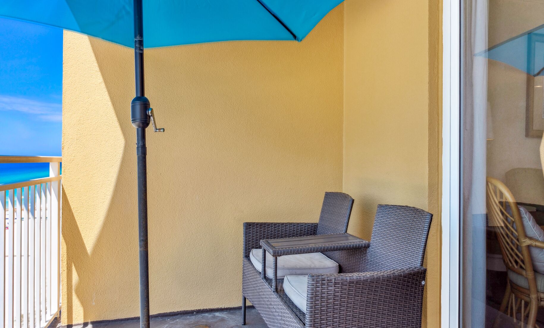A balcony with a wicker chair and a table, a blue umbrella, and a view of the ocean.