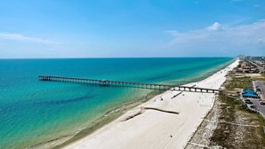 Ein Küstenstrand mit einem Steg, der ins Meer führt, einer sandigen Uferlinie und einer Stadtlandschaft im Hintergrund.